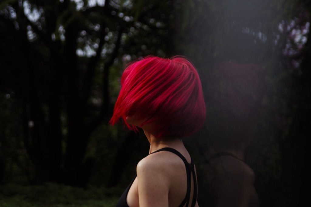 Woman with vibrant red hair turns away in a shadowy forest, striking dynamic pose in natural setting.
